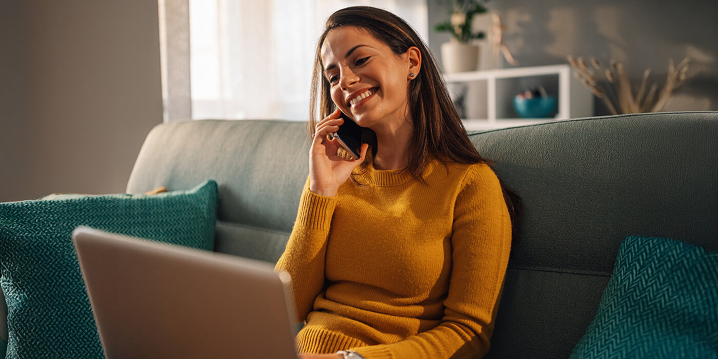 Eine junge Frau telefoniert entspannt von ihrem Sofa aus, während sie gleichzeitig am Laptop arbeitet. Das Bild steht für eine zuverlässige Telefonverbindung im Alltag – komfortabel, klar und flexibel über den Glasfaseranschluss.