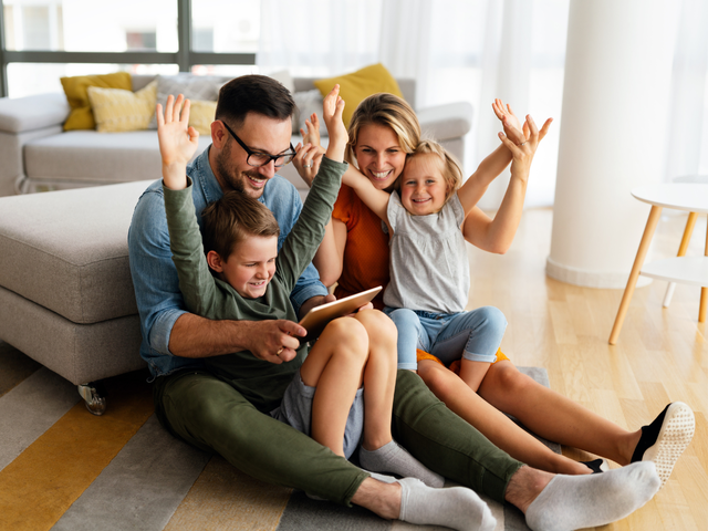 Familie sitzt gemeinsam im Wohnzimmer und nutzt ein Tablet, während Eltern und Kinder fröhlich die Hände heben.
