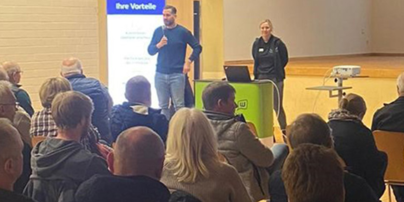 Viele Personen sitzen in einem Saal, Projektverantwortliche stehen vorne mit Mikrofon