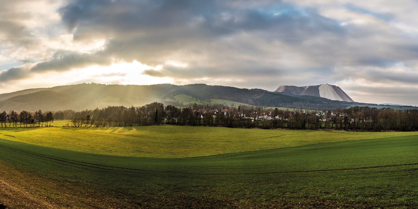 Weite Landschaftsaufnahme von Heringen mit grünem Feld, angrenzendem Wald und dem markanten Kaliberg bei bewölktem Himmel – Sinnbild für moderne Infrastruktur im ländlichen Raum.