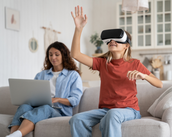 Frau sitzt mit Laptop auf dem Schoß auf dem Sofa, daneben ein Mädchen mit einer VR Brille