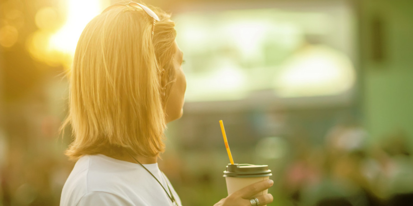 Frau mit Trinkbecher in der Hand steht vor einer großen Fernsehleinwand