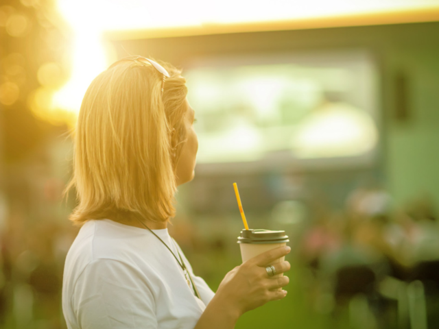 Frau mit Trinkbecher in der Hand steht vor einer großen Fernsehleinwand