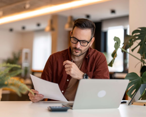 Mann sitzt am Schreibtisch vor dem Laptop und hält ein Dokument in der Hand