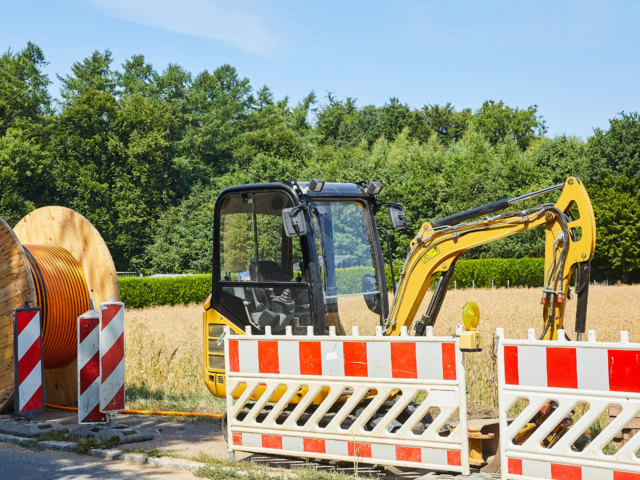 Bagger steht am Straßenrand, ein Feld ist im Hintergrund